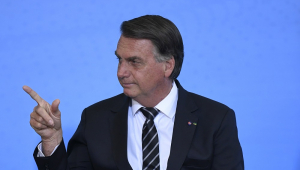 Sob um fundo azul, vestido de terno e gravata, mas sem máscara, Bolsonaro inclina a cabeça para a direita e aponta com o indicador estendido e o polegar levantado