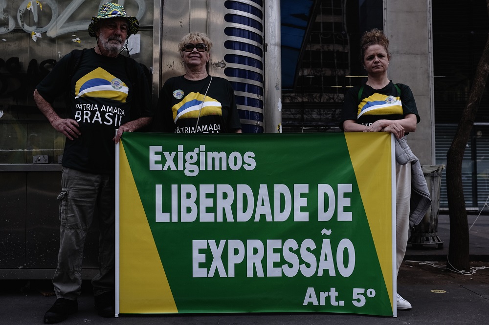Três pessoas (um homem e duas mulheres), usando a mesma camisa preta com a mensagem Pátria Amada Brasil, posam à frente de um cartaz em verde e amarelo, com a frase 