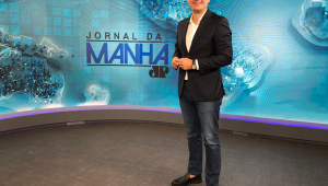 O jornalista Bruno Meyer em pé no estúdio do Jornal da Manhã, com o logo do telejornal ao fundo, vestido com calça jeans, paletó, camisa e sapato, usando um relógio no pulso esquerdo