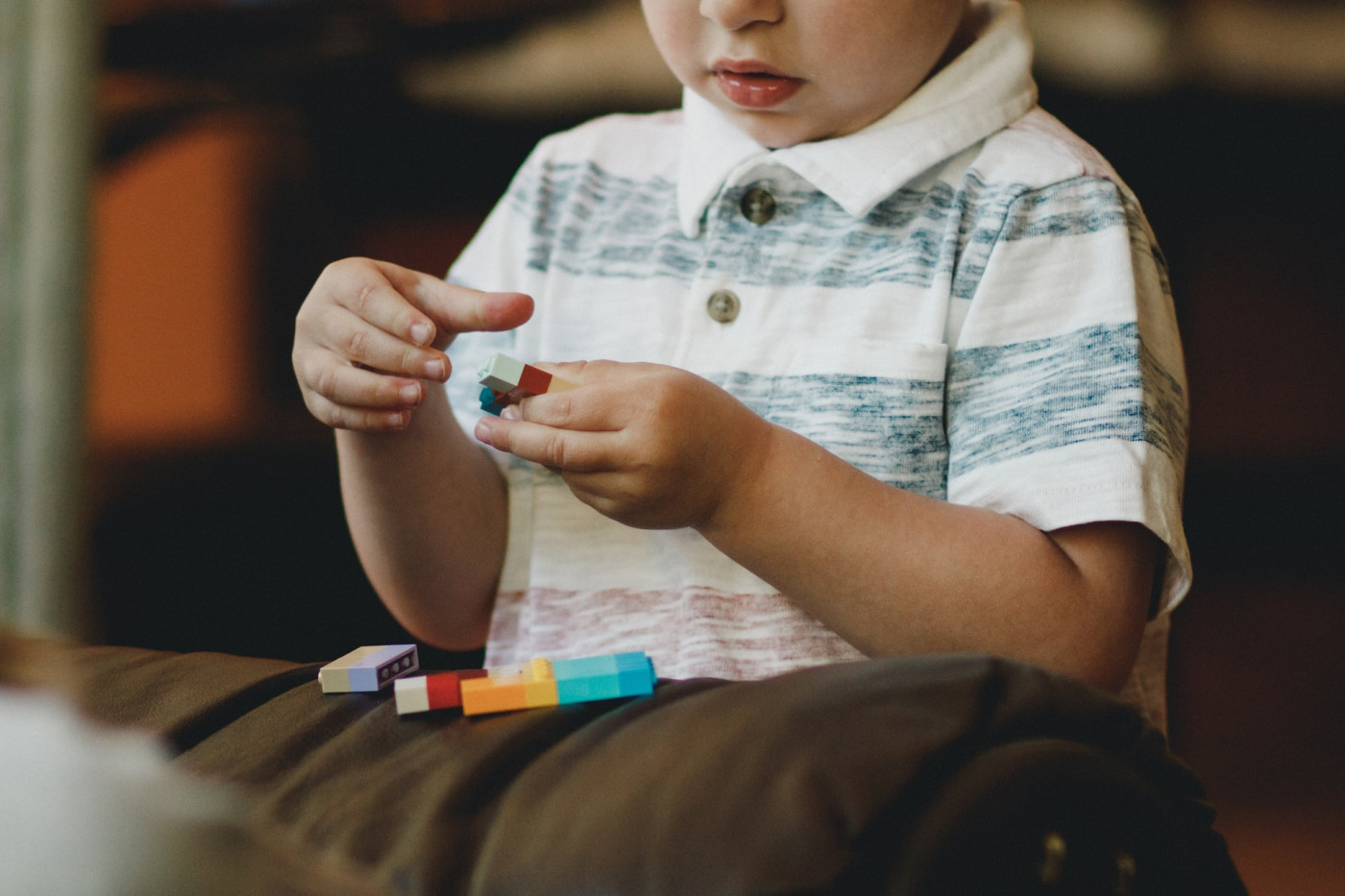 Criança brincando com blocos coloridos em cima de um sofá marrom. Só aparece metade do rosto e uma camiseta pólo branca com listras verdes e vermelhas