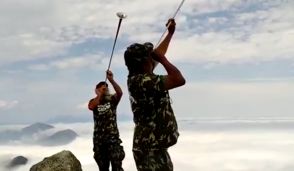 Dois religiosos com blusas camufladas tocando trombeta no Pico do Dedo de Deus, no Rio de Janeiro