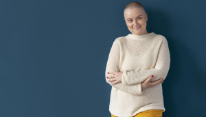 Mulher em tratamento contra o câncer, de braços cruzados, parece confiante diante de uma parede roxa