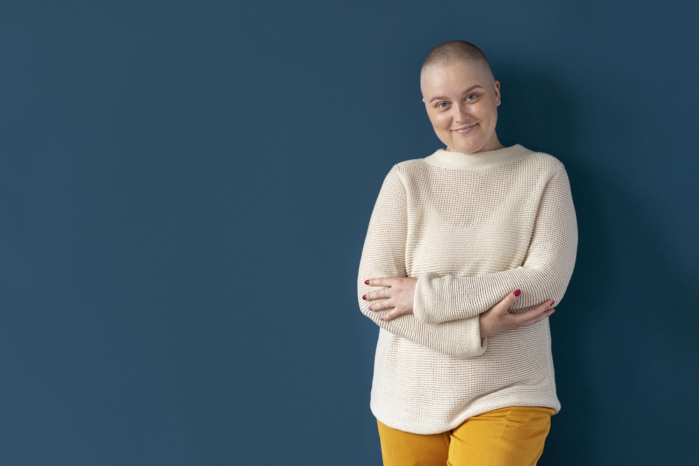 Mulher em tratamento contra o câncer, de braços cruzados, parece confiante diante de uma parede roxa
