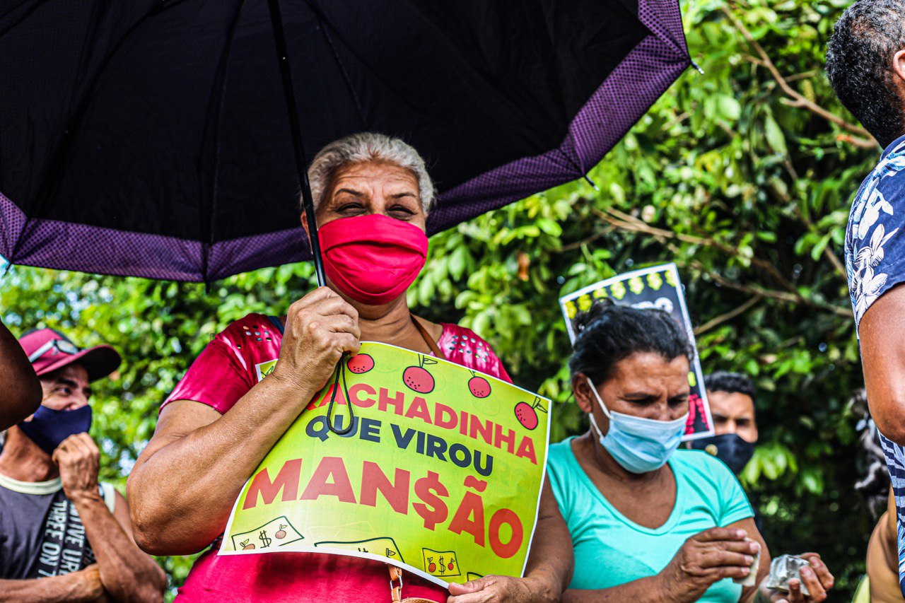 Senhora segurando um cartaz escrito 'Rachadinha que virou mansão' em frente à mansão de Flávio Bolsonaro