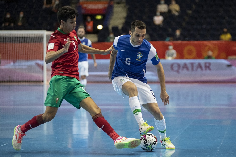 Marcado por marroquino (de camisa vermelha e shorts verde), Jogador da seleção brasileira de futsal (de camisa azul e shorts branco) pisa na bola e tenta protegê-la