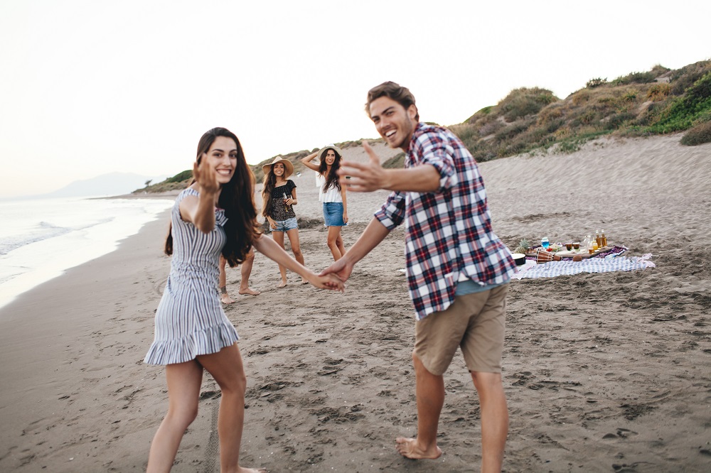 Casal de jovens brancos atraentes de mãos dadas olha para a câmera e faz com as mãos um convite para a festa com amigos que está acontecendo na areia da praia