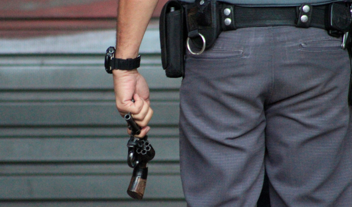 Policial militar aparece de costas segurando uma arma