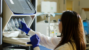 Uma cientista com jaleco branco e luvas azuis manuseia com a mão direita experimento de sequenciamento de genoma