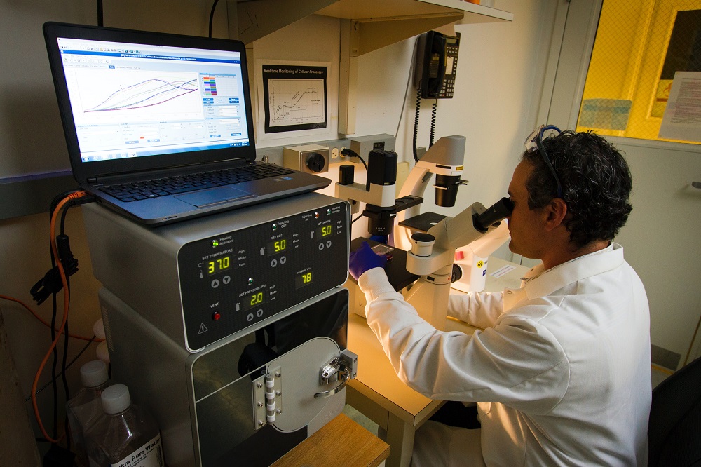 Dentro de um laboratório, médico, vestido com um jaleco branco, olha pela lente de um microscópio para observar resultados de um exame, à sua esquerda, um computado, em altura superior à cabeça do doutor, mostra gráficos