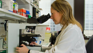 Uma cientista loira, de jaleco branco, observa pelo microscópio em uma sala