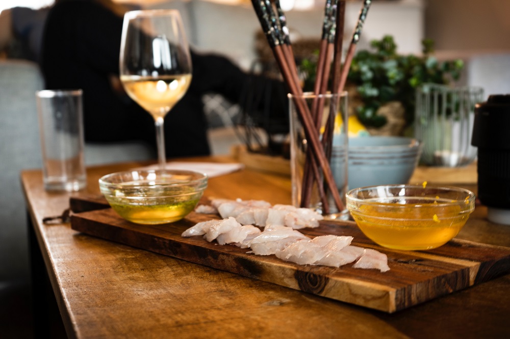Fatias de peixe cru sobre uma tpábua posta sobre uma mesa, com uma taça de vinho branco ao lado