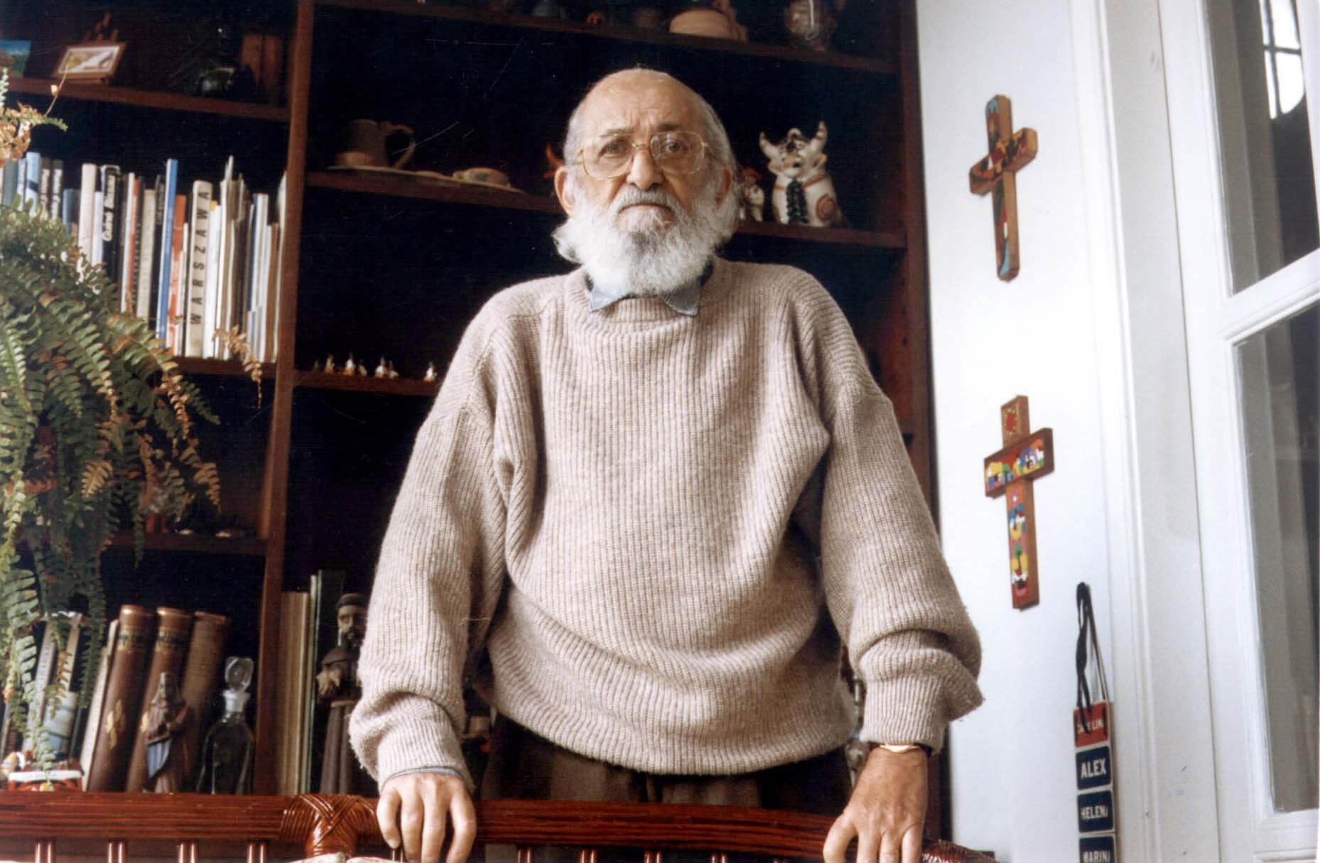 Paulo Freire com suéter claro, em pé, apoiado numa cadeira olhando para a câmera