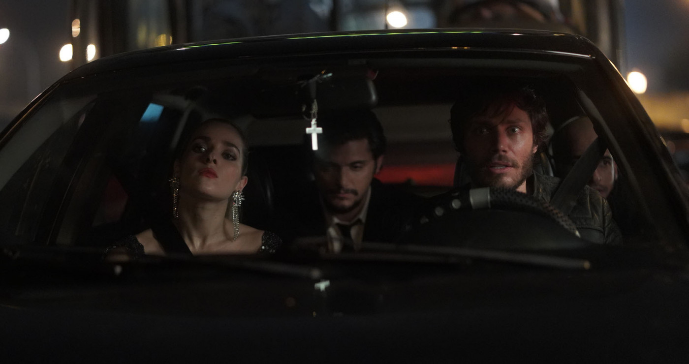 Três pessoas dentro de um carro com um cenário escuro. O motorista é um homem, tem uma mulher sentada ao lado e atrás outro homem. Um crucifixo está pendurado