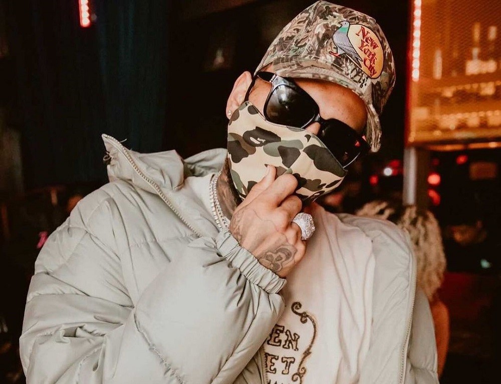 De máscara camuflada, boné, óculos escuros e uma jaqueta cinza clara, Chris Brown inclina a cabeça e coloca a mão direita no queixo