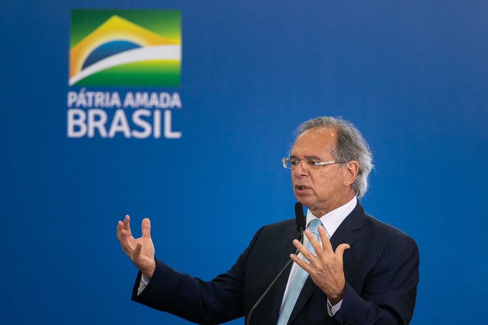Em trajes sociais, mas sem gravata, Paulo Guedes abre os braços enquanto fala; atrás dele, uma parede azul com a bandeira do país estilizada e a frase Pátria Amada Brasil
