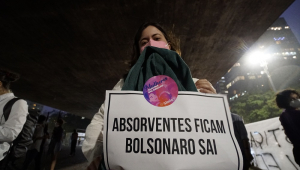 No vão livre do Masp, Samia Bonfim posa de máscara com um cartaz escrito "absorventes ficam, Bolsonaro sai", colado à roupa dela com um adesivo com a mensagem "Mulheres contra Bolsonaro"