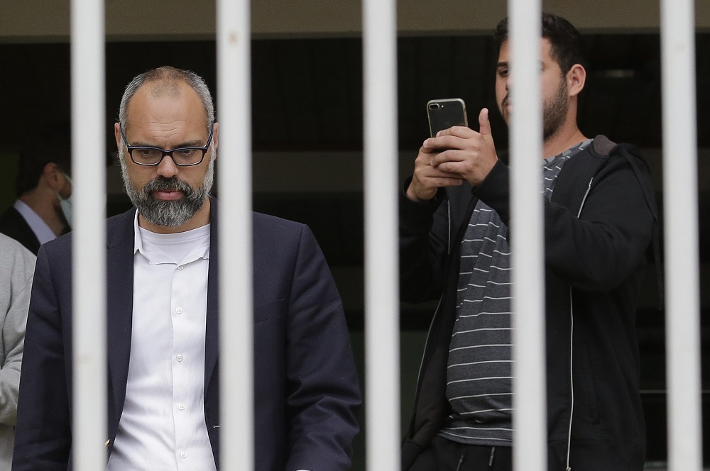 O jornalista Allan dos Santos à frente de um portão, que, devido ao enquadramento da foto, faz parecer que ele está atrás das grades