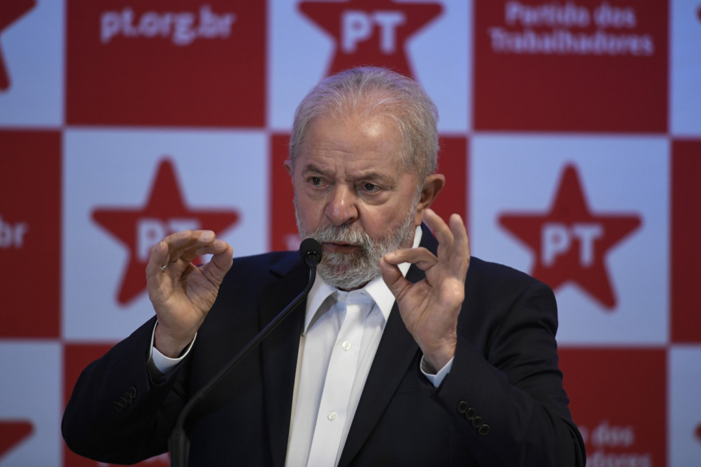 Presidente Lula unindo os dois polegares aos indicadores e falando em um microfone. Usa terno e camisa branca. Atrás, logos do PT