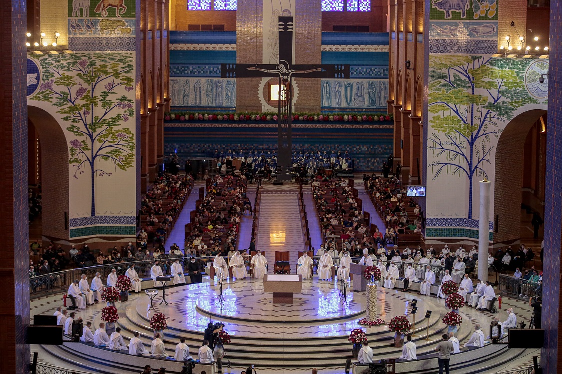 Imagem de uma igreja em Aparecida com uma espécie de elevado redondo e vários padres sentados em cadeira em uma roda. No meio, um altar. Atrás, há duas fileiras com várias pessoas sentadas em bancos de igreja.