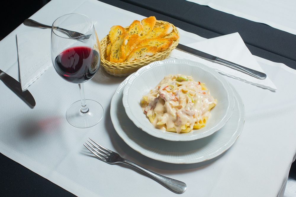 Prato de Penne com milho branco e vegetais, colocado à mesa com um copo de vinho e uma porção de torradas