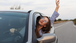 Jovem mulher de óculos dirige carro prata na estrada, à luz do dia, e coloca a cabeça e o braço esquerdo pra fora