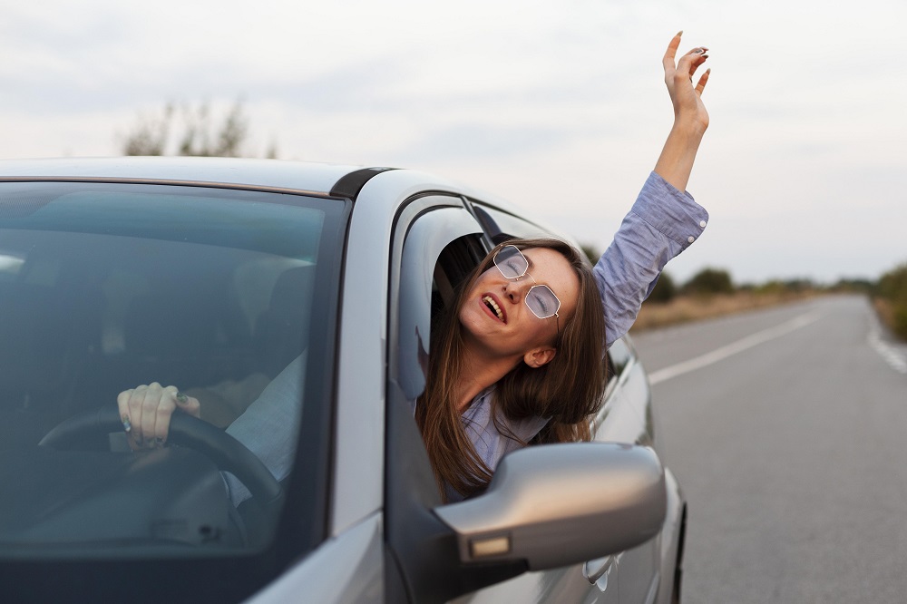 Jovem mulher de óculos dirige carro prata na estrada, à luz do dia, e coloca a cabeça e o braço esquerdo pra fora