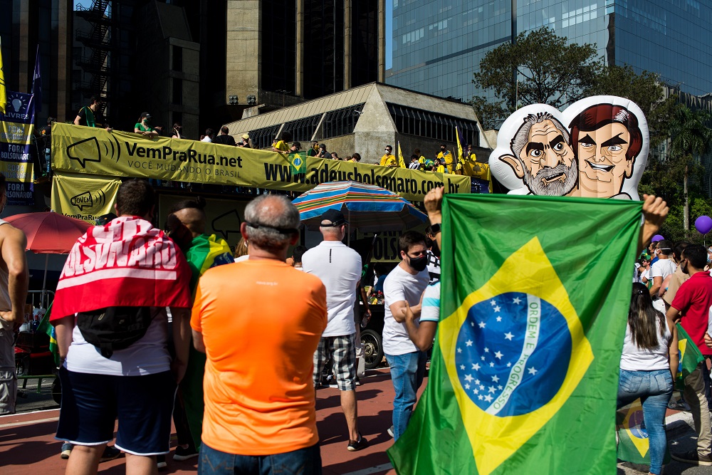 Boneco do Lula e de Bolsonaro juntos, parcialmente encobertos pela bandeira do Brasil, durante manifestação contra Bolsonaro na avenida Paulista