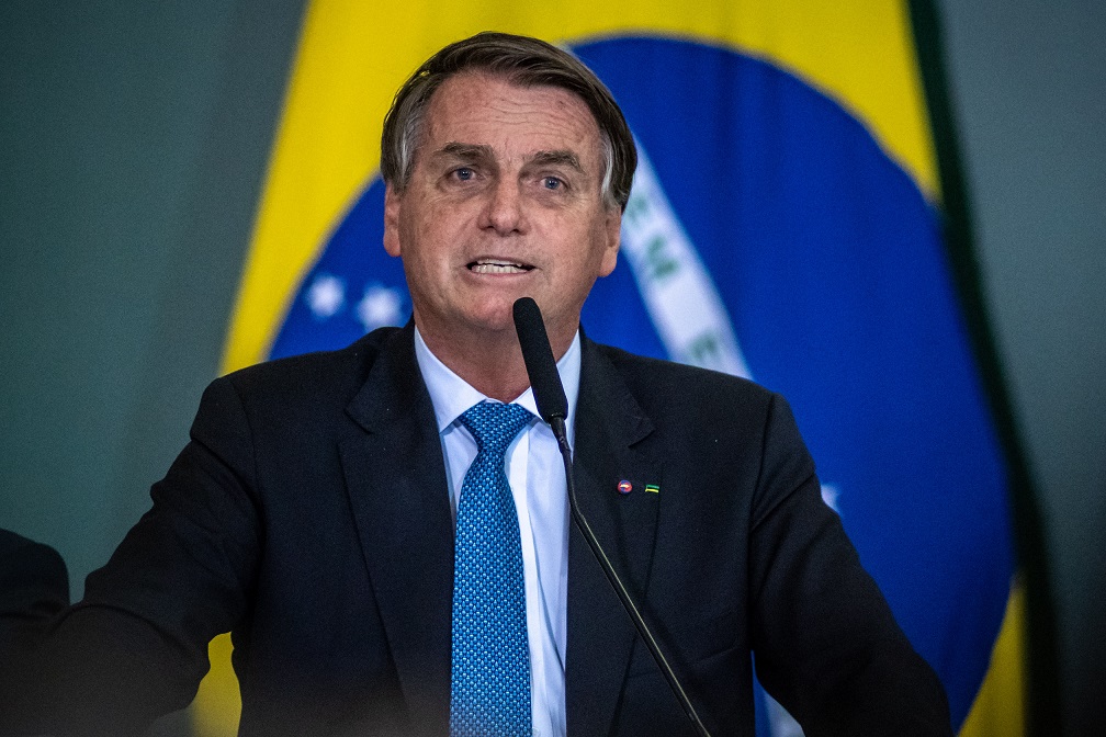 O presidente Jair Bolsonaro falando em microfone com uma bandeira do Brasil ao fundo