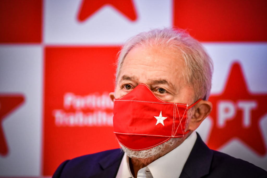 Ex-presidente Lula olhando para o horizonte com uma máscara de proteção vermelha com uma estrela branca. Atrás, logos do PT