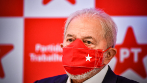 Ex-presidente Lula olhando para o horizonte com uma máscara de proteção vermelha com uma estrela branca. Atrás, logos do PT