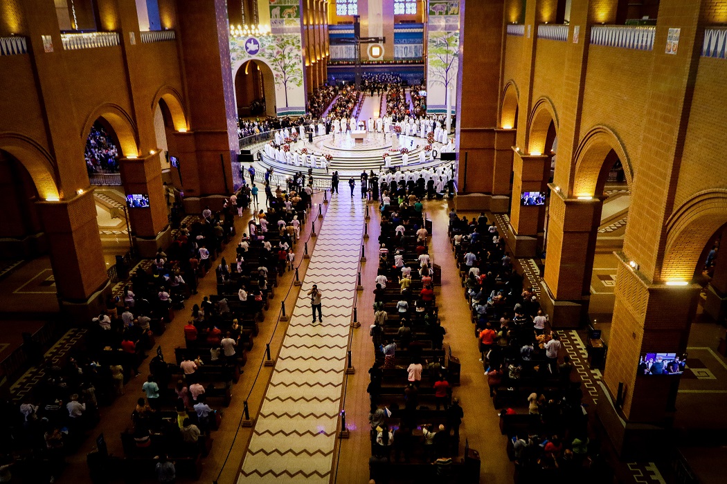Foto tirada do alto e quatro fileiras com várias pessoas sentadas em cadeiras em uma igreja. No meio, há um caminho branco.