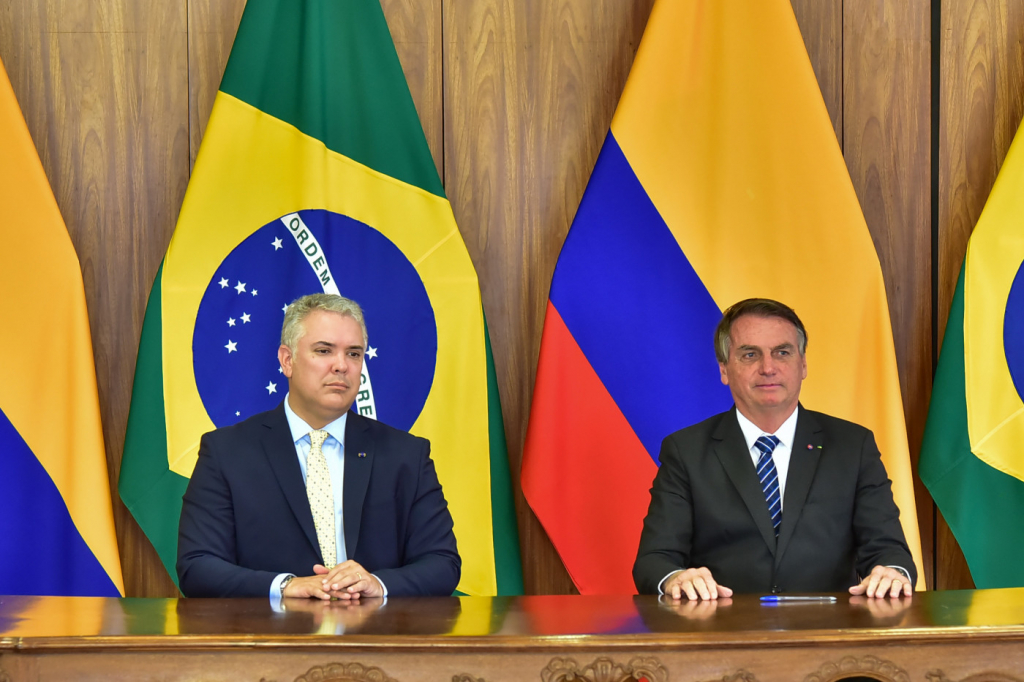O presidente Ivan Duque, da Colômbia, e presidente Jair Bolsonaro sentados em uma bancada em frente às bandeiras dos países