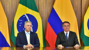 O presidente Ivan Duque, da Colômbia, e presidente Jair Bolsonaro sentados em uma bancada em frente às bandeiras dos países