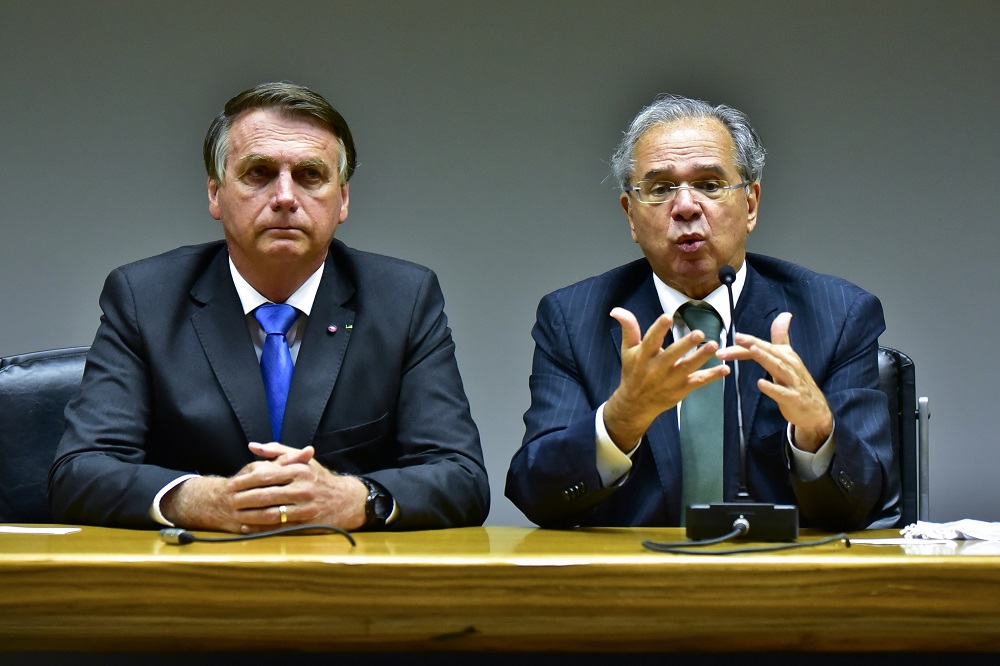 Jair Bolsonaro e Paulo Guedes lado a lado, sentados à frente de uma mes
