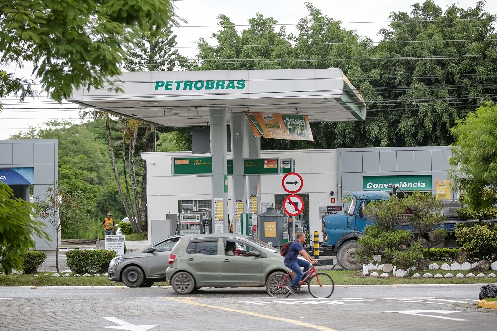 Posto de gasolina da Petrobras com um carro parado para abastecer; na rua, um carro popular e um ciclista passam em frente ao estabelecimento