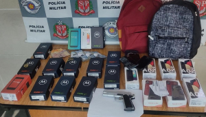 Mesa com diversas caixas de celulares dispostas lado a lado, duas mochilas no fundo e uma pistola; no fundo, um banner da Polícia Militar