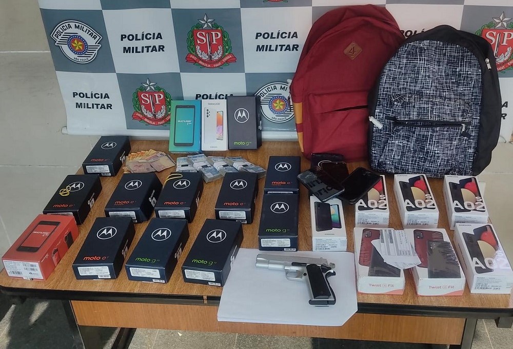 Mesa com diversas caixas de celulares dispostas lado a lado, duas mochilas no fundo e uma pistola; no fundo, um banner da Polícia Militar