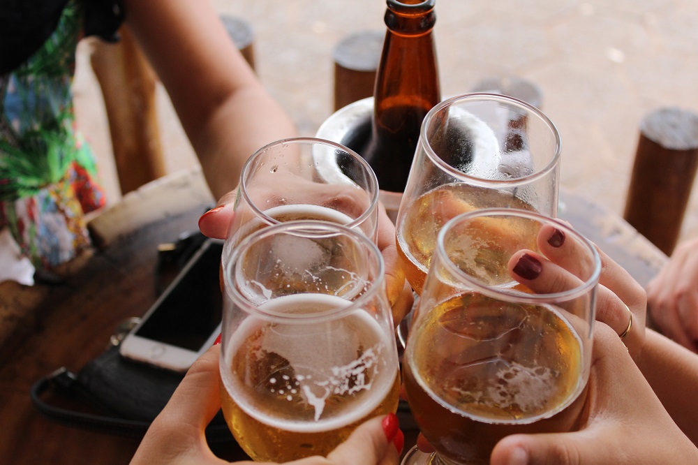 Imagem de um brinde com quatro copos de cerveja, em que a garrafa, com um isolante térmico, aparece sobre a mesa