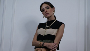 Fernanda Perlaky Derenze, uma mulher morena, na faixa dos 20/30 anos, usa blusinha sem manga com listras horizontais, colar, brincos e posa para foto de braços cruzados