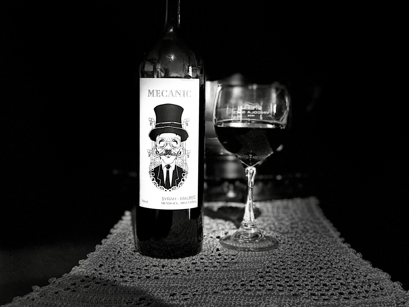 Foto em preto e branco de uma vinho com rótulo de um homem usando chapéu e uma taça servida pela metade ao lado