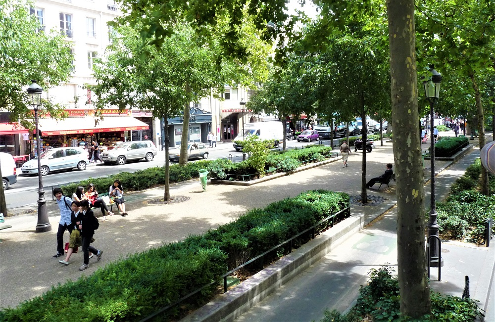 Rua com duas calçadas, sendo uma elas bem ampla, com bancos, alguns transeuntes, jardins e diversas árvores