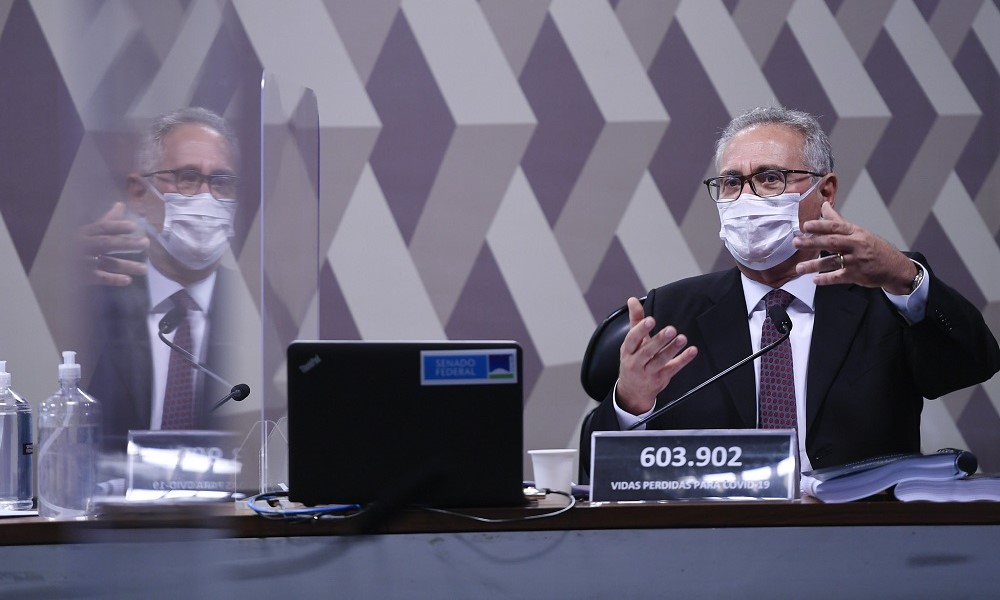 Renan Calheiros, em trajes sociais e máscara, gesticula durante pronunciamento em sua cabine na CPI da Covid-19, com sua imagem refletida pela divisória transparente