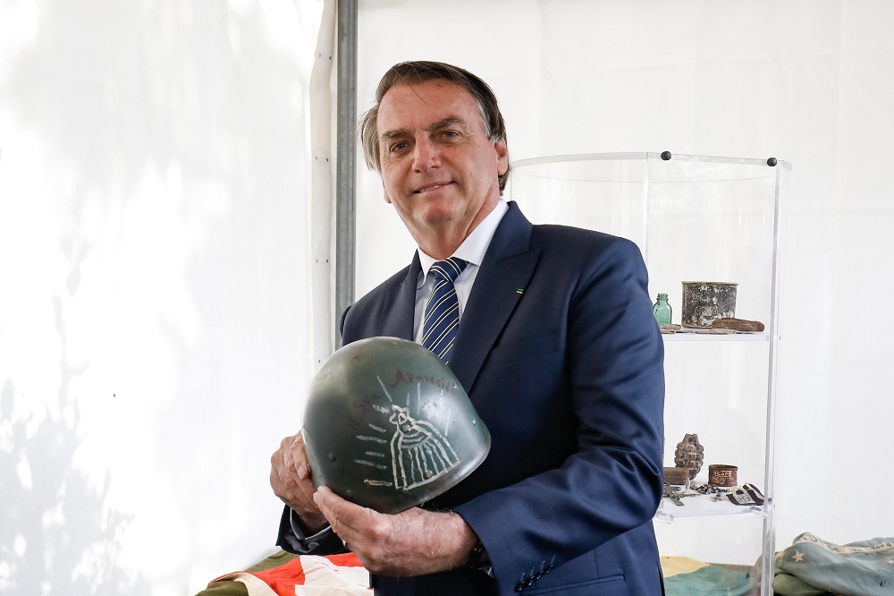 De terno e gravata, sorrindo, o presidente Jair Bolsonaro segura um capacete da Segunda Guerra