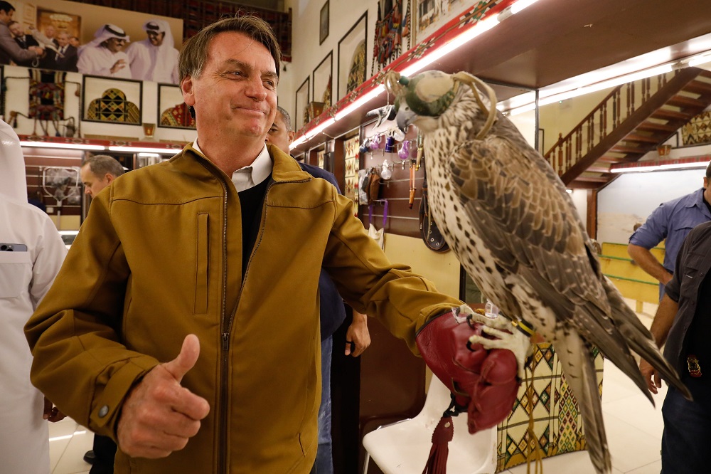 Equilibrando uma águia de enfeite na mão esquerda, Bolsonaro sorri dentro de um mercado árabe cheio de produtos típicos