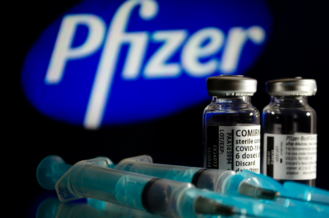 Dois frascos de vacina e duas seringas; Ao fundo, um logo da Pfizer