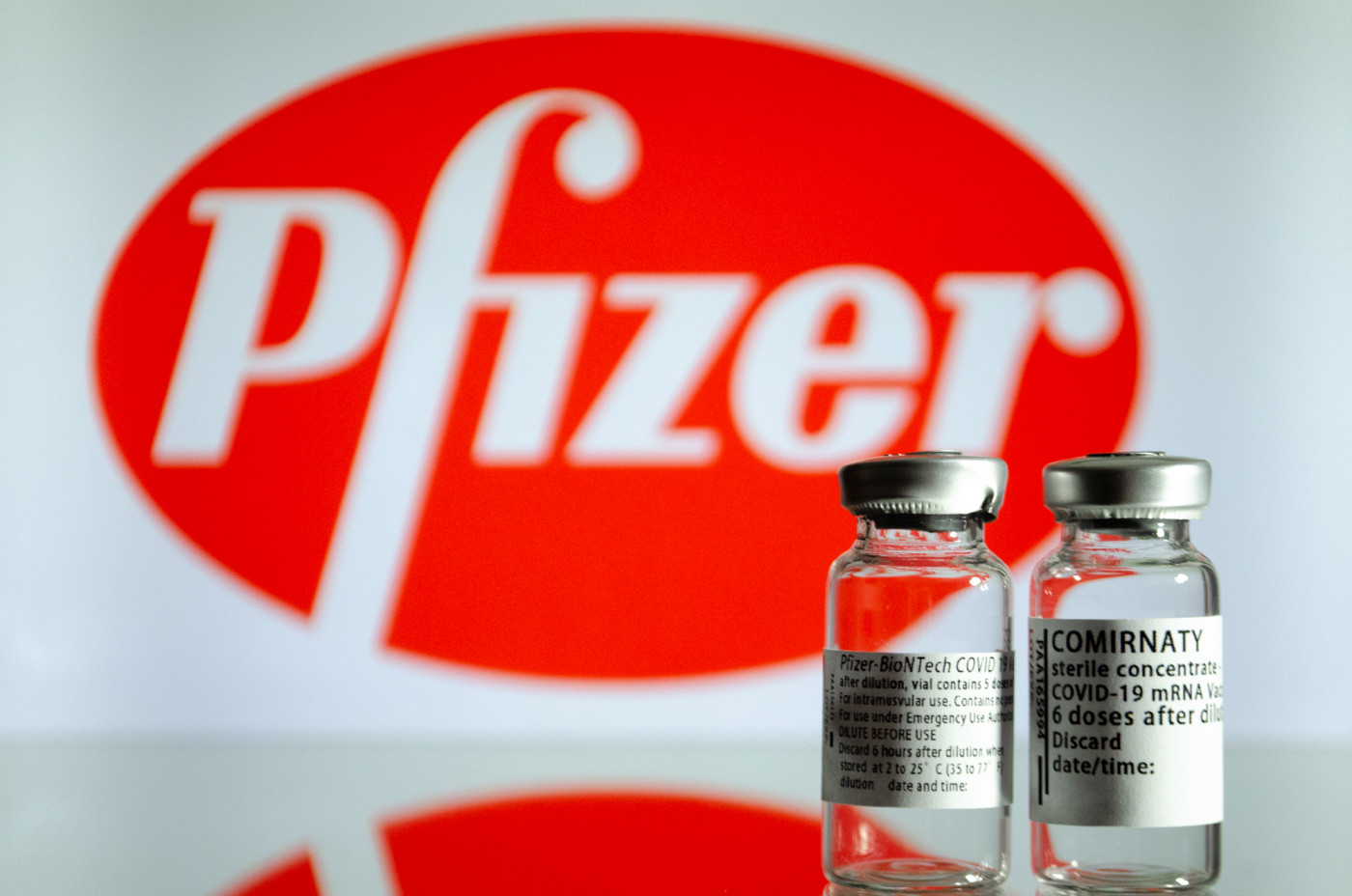 Dois frascos de vacina contra a Covid-19 da Pfizer; ao fundo, o logo da farmacêutica