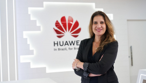 Mulher branca na faixa dos 40 anos de roupa executiva preta e braços cruzados em frente a uma parede de vidro com o símbolo da Huawei