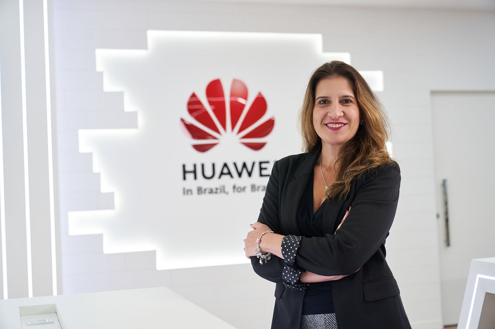 Mulher branca na faixa dos 40 anos de roupa executiva preta e braços cruzados em frente a uma parede de vidro com o símbolo da Huawei