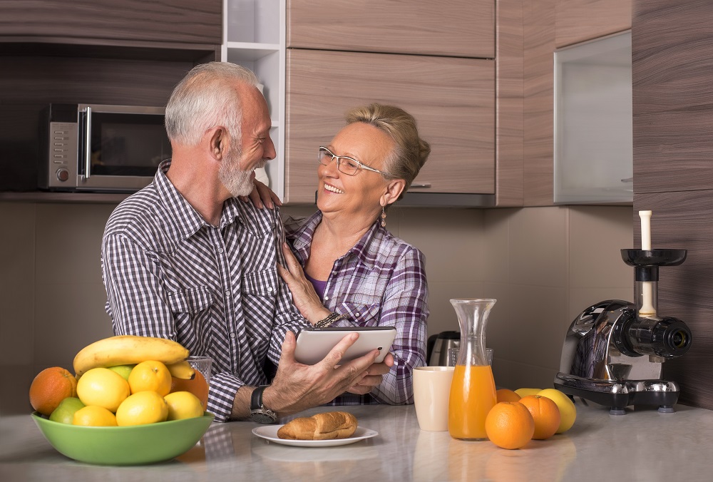 Casal de idosos brancos se entreolham e sorriem na cozinha debruçados na mesa, que tem uma cesta com bananas, uma baguete no prato, uma jarra de suco de laranja, três laranjas e um copo plástico; ele segura um tabelt, e ela coloca as mãos nos ombros dele