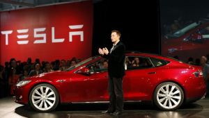 Elon Musk, de roupa esporte fino, em pé ao lado de carro vermelho da Tesla durante evento da montadora